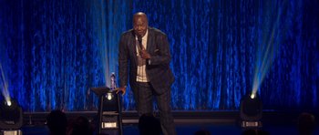 Movie still from “Chappelle's Home Team - Earthquake: Legendary” (2022), directed by Stan Lathan – A man standing on a stage holding a microphone; Medium shot, High angle