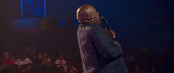 Movie still from “Chappelle's Home Team - Earthquake: Legendary” (2022), directed by Stan Lathan – A man holding a microphone while standing on a stage; Medium shot, Over the shoulder angle