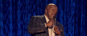 Movie still from “Chappelle's Home Team - Earthquake: Legendary” (2022), directed by Stan Lathan – A man in a suit and tie holding a microphone and singing; Close Up shot, High angle