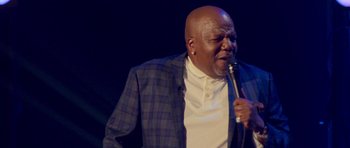 Movie still from “Chappelle's Home Team - Earthquake: Legendary” (2022), directed by Stan Lathan – A man in a blue and white jacket holding a microphone; Close Up shot, Low angle