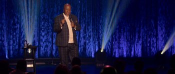 Movie still from “Chappelle's Home Team - Earthquake: Legendary” (2022), directed by Stan Lathan – A man standing on a stage holding a microphone; Medium shot, Low angle