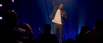 Movie still from “Chappelle's Home Team - Earthquake: Legendary” (2022), directed by Stan Lathan – A man standing on a stage holding a microphone; Wide shot, Low angle