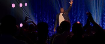 Movie still from “Chappelle's Home Team - Earthquake: Legendary” (2022), directed by Stan Lathan – A man in a suit and tie on a stage waving; Medium shot, Low angle