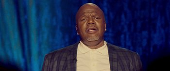 Movie still from “Chappelle's Home Team - Earthquake: Legendary” (2022), directed by Stan Lathan – A man with a beard and a jacket is looking at the camera; Close Up shot, Low angle