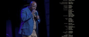 Movie still from “Chappelle's Home Team - Earthquake: Legendary” (2022), directed by Stan Lathan – A man in a blue jacket is holding a microphone; Medium shot, Low angle