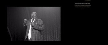 Movie still from “Chappelle's Home Team - Earthquake: Legendary” (2022), directed by Stan Lathan – A black and white photo of a man in a suit holding a microphone; Medium shot, High angle
