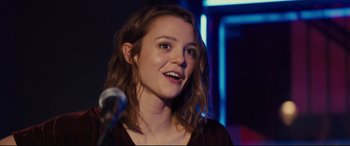 Movie still from “A Dog's Journey” (2019), directed by Gail Mancuso – A young woman smiles while standing in front of a microphone; Close Up shot, Over the shoulder angle