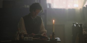 Movie still from “Ehrengard: The Art of Seduction” (2023), directed by Bille August – A man sitting at a table reading a book by a lit candle; Medium shot, Over the shoulder angle