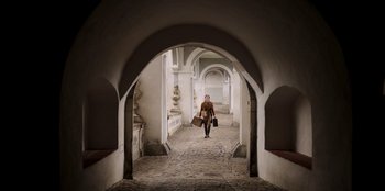 Movie still from “Ehrengard: The Art of Seduction” (2023), directed by Bille August – A woman walking down an arched walkway holding two suitcases; Wide shot, Low angle