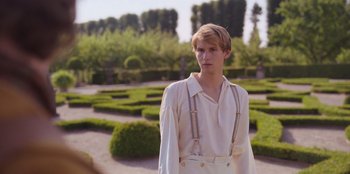 Movie still from “Ehrengard: The Art of Seduction” (2023), directed by Bille August – A young man wearing suspenders standing next to a garden; Medium shot, Over the shoulder angle