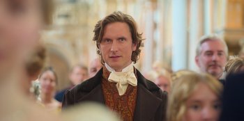 Movie still from “Ehrengard: The Art of Seduction” (2023), directed by Bille August – A man with long brown hair wearing a suit and bow tie; Close Up shot, Over the shoulder angle
