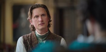 Movie still from “Ehrengard: The Art of Seduction” (2023), directed by Bille August – A man with long blonde hair wearing a vest and neck tie; Close Up shot, Over the shoulder angle