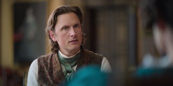 Movie still from “Ehrengard: The Art of Seduction” (2023), directed by Bille August – A man with long blonde hair wearing a vest and tie; Close Up shot, Over the shoulder angle