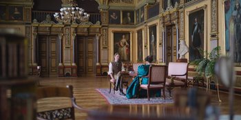 Movie still from “Ehrengard: The Art of Seduction” (2023), directed by Bille August – Two people are sitting in a room with paintings on the walls; Wide shot, High angle