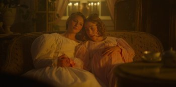 Movie still from “Ehrengard: The Art of Seduction” (2023), directed by Bille August – Two young girls are sitting on a couch together; Medium shot, High angle