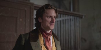 Movie still from “Ehrengard: The Art of Seduction” (2023), directed by Bille August – A man with long brown hair wearing a green jacket and a red neck tie; Close Up shot, Over the shoulder angle