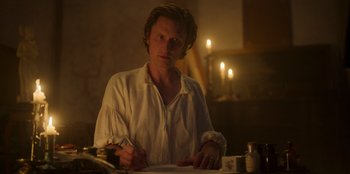 Movie still from “Ehrengard: The Art of Seduction” (2023), directed by Bille August – A man sitting at a table writing on a piece of paper; Medium shot, Over the shoulder angle