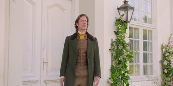 Movie still from “Ehrengard: The Art of Seduction” (2023), directed by Bille August – A man standing in front of a building wearing a green coat; Medium shot, Low angle