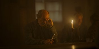 Movie still from “Ehrengard: The Art of Seduction” (2023), directed by Bille August – An older man sitting at a table with a candle in front of him; Close Up shot, Over the shoulder angle