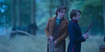 Movie still from “Ehrengard: The Art of Seduction” (2023), directed by Bille August – A man holding a walking stick next to another man in a forest; Medium shot, Over the shoulder angle