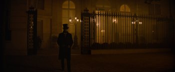 Movie still from “Eiffel” (2021), directed by Martin Bourboulon – A man in a top hat and a long coat standing in front of an iron gate; Wide shot, Over the shoulder angle