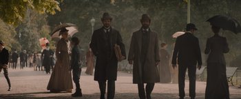 Movie still from “Eiffel” (2021), directed by Martin Bourboulon – A couple of men walking down a street holding umbrellas; Wide shot, Over the shoulder angle