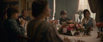 Movie still from “Eiffel” (2021), directed by Martin Bourboulon – A woman sitting at a dinner table with a flower arrangement in front of her; Medium shot, Over the shoulder angle