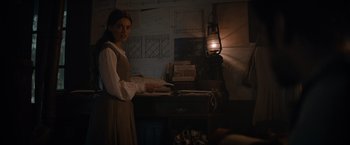 Movie still from “Eiffel” (2021), directed by Martin Bourboulon – A woman standing in front of a table in a dimly - lit room; Medium shot, Over the shoulder angle
