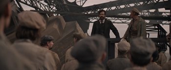 Movie still from “Eiffel” (2021), directed by Martin Bourboulon – A man standing in front of a group of people; Wide shot, Over the shoulder angle