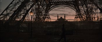 Movie still from “Eiffel” (2021), directed by Martin Bourboulon – A person walking under an arch near a fence; Extreme Wide shot, Low angle