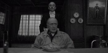 Movie still from “El Conde” (2023), directed by Pablo Larraín – A man sitting at a table with another man standing behind him; Medium shot, Low angle