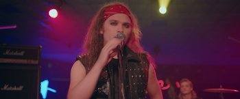 A man with long curly hair wearing a red bandanna is holding a microphone