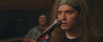 A man with long dreadlocks wearing a headband is speaking into a microphone