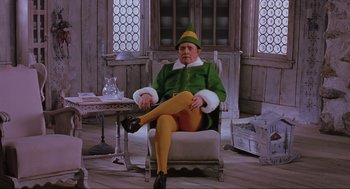 Movie still from “Elf” (2003), directed by Jon Favreau – A man dressed as the elf sitting in a chair; Wide shot, Low angle