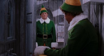 Movie still from “Elf” (2003), directed by Jon Favreau – A man dressed up as buddy the elf standing next to another man; Medium shot, Over the shoulder angle