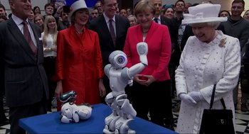Movie still from “Elizabeth: A Portrait in Part(s)” (2022), directed by Roger Michell – A group of people standing next to a table with a robot; Wide shot, Over the shoulder angle
