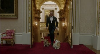 Movie still from “Elizabeth: A Portrait in Part(s)” (2022), directed by Roger Michell – A man in a tuxedo walking down the stairs with two small dogs; Wide shot, Low angle