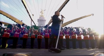Movie still from “Elizabeth: A Portrait in Part(s)” (2022), directed by Roger Michell – A man holding a microphone while standing in front of a group of people; Extreme Wide shot, Low angle