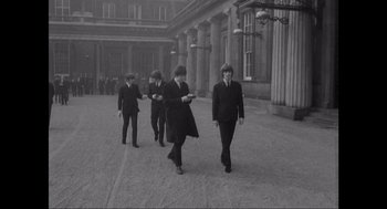 Movie still from “Elizabeth: A Portrait in Part(s)” (2022), directed by Roger Michell – A black and white photo of a group of men walking down a street; Wide shot, High angle