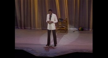 Movie still from “Elizabeth: A Portrait in Part(s)” (2022), directed by Roger Michell – A man standing on a stage holding a microphone; Wide shot, High angle