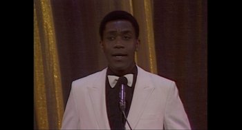 Movie still from “Elizabeth: A Portrait in Part(s)” (2022), directed by Roger Michell – A man in a white suit is speaking at a microphone; Medium shot, High angle