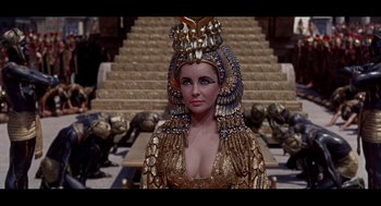 Movie still from “Elizabeth: A Portrait in Part(s)” (2022), directed by Roger Michell – A woman wearing a gold dress and a crown; Close Up shot, High angle