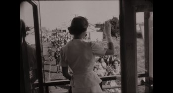 Movie still from “Elizabeth: A Portrait in Part(s)” (2022), directed by Roger Michell – An old black and white photo of a woman waving at a crowd of onlookers; Medium shot, Over the shoulder angle