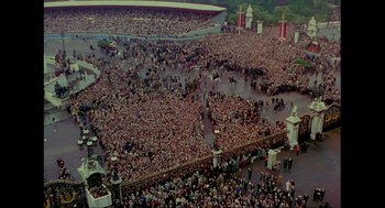 Movie still from “Elizabeth: A Portrait in Part(s)” (2022), directed by Roger Michell – An aerial view of a large crowd of people in a stadium; Extreme Wide shot, High angle