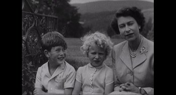 Movie still from “Elizabeth: A Portrait in Part(s)” (2022), directed by Roger Michell – An older woman and two young boys sitting next to each other in a field; Medium shot, High angle