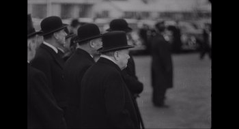Movie still from “Elizabeth: A Portrait in Part(s)” (2022), directed by Roger Michell – A black and white photo of a group of men in suits and top hats; Medium shot, Low angle
