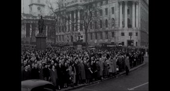 Movie still from “Elizabeth: A Portrait in Part(s)” (2022), directed by Roger Michell – A large group of people standing on the side of the street; Extreme Wide shot, High angle