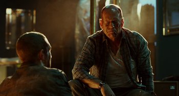 Movie still from “A Good Day to Die Hard” (2013), directed by John Moore – A man sitting next to another man in a dimly - lit room; Close Up shot, Over the shoulder angle