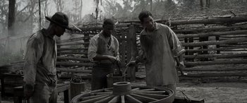 Movie still from “Emancipation” (2022), directed by Antoine Fuqua – Two men working on a wheel in an outdoor setting; Medium shot, Low angle