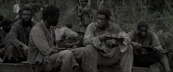 Movie still from “Emancipation” (2022), directed by Antoine Fuqua – A group of men sitting next to each other in the woods; Medium shot, Over the shoulder angle